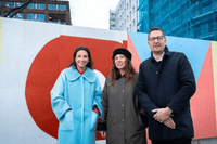 Tre personer, två kvinnor och en man, står framför ett byggplank målat med stora, färgglada geometriska former i rött, orange, rosa och gult. Kvinnan till vänster bär en ljusblå kappa, kvinnan i mitten bär en mörk kappa och basker, och mannen till höger b