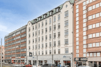 En ljusgul, sekelskiftesliknande flervåningsbyggnad placerad mellan två mer moderna kontorshus i tegel och glas längs en stadsilag.