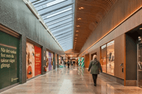En ljus och modern galleriagång med glastak och träpaneler i taket. Till vänster syns stora reklamaffischer och till höger butiksfönster. Enstaka människor promenerar genom lokalen.