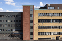 En stor kontors- eller industribyggnad med tre tydligt olika fasader: en mörkgrå sektion till vänster, en vertikal del i rött tegel i mitten och en gul tegelvägg med rader av fönster till höger. Byggnaden står under en klarblå himmel med vita moln