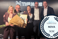Fem glada personer poserar tillsammans på en scen framför skylten "THE NODE". De håller i en stor blombukett och ett inramat diplom. I bildens högra hörn finns ett emblem som visar att köpcentrumet NOVA har vunnit "Commercial Space Shopping Center" vid NC
