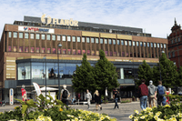 Exteriör av gallerian Flanör i Gävle sett från Stortorget. Den bruna tegelbyggnaden har stora glaspartier på gatuplan, skyltar för butiker som Intersport och en takskylt med namnet Flanör. Människor promenerar på det stenlagda torget framför gula blommor 