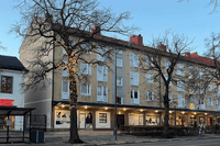 Bild tagen under kvällstid av ett bostadshus med tre våningar i ljusgul puts. På gatuplanet finns flera butiker under ett utskjutande tak med inbyggd belysning. Flera kala träd står framför huset, varav ett är dekorerat med julbelysning