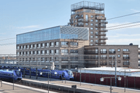 Blå pendeltåg vid en tågstation framför en modern tegelbyggnad med stora glaspartier och en hög huskropp.