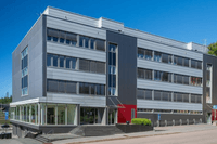 Modern kontorsbyggnad i fyra våningar med grå fasadpaneler, röda accentdetaljer och entrétrappa mot gatan under en klarblå sky.