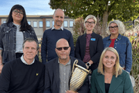 En grupp på sju personer poserar utomhus framför buskar och träd. Tre personer sitter på en bänk i främre raden, varav en man i mitten håller i en stor pokal. Fyra personer står bakom dem. Alla ler eller har ett neutralt uttryck, och de är klädda i vardag