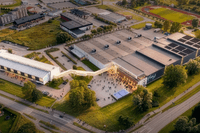 En flygbild över Plejds huvudkontor och produktionsanläggning i Mölndal. Den moderna industrifastigheten har stora lagerhallar och en vit kontorsbyggnad som är sammanlänkad via en inglasad gångbro. Framför byggnaden syns en öppen samlingsplats med människ