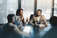 Fyra affärsklädda personer sitter i ett möte runt ett bord i ett ljust konferensrum med stora fönster. En kvinna i mitten talar engagerat och gestikulerar, medan de övriga lyssnar uppmärksamt. På bordet syns bärbara datorer och surfplattor.