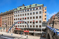 Exteriörbild i fågelperspektiv över ett stadslandskap med flervåningsbyggnader, biografen Saga, en vägbro och en blomsterbåge under klarblå sky.
