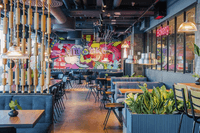 Interiör av ett modernt och färgstarkt café eller bageri. Lokalen har en stor popkonst-väggmålning med texten "MR CAKE SWEET", blå soffgrupper, träbord och en unik rumsavdelare gjord av kavel-liknande trädetaljer. Stora glasfönster ger inblick till ett ba