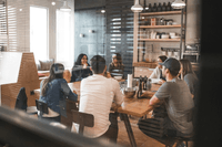 En grupp människor sitter samlade runt ett långt träbord i en avslappnad kontorsmiljö eller fikarum. De samtalar i ett ljust rum med industriell känsla, där en öppen hylla med porslin och stora fönster syns i bakgrunden.