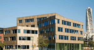 Ett modernt kontorshus i flera nivåer med stora glasfönster, beläget i Västra Hamnen i Malmö. I bakgrunden syns det ikoniska Turning Torso resa sig mot den blå himlen. I förgrunden syns grönska, parkeringsplatser och en vit bil parkerad längs gatan.