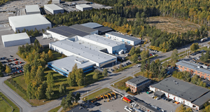Flygfoto över ett industriområde med stora lagerbyggnader och verkstäder omgivna av skog och parkeringsplatser i höstfärger.