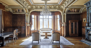 En praktfull och detaljrik interiör i klassisk stil med väggar och tak i snidat ljust trä. Rummet har en stor kristallkrona, mönstrat parkettgolv och ett välvt tak med dekorativa målningar. Två vita stolar och ett litet bord står placerade mot den bakre v