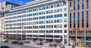 Byggnaden är en stor kontors- och affärsfastighet med en ljus fasad, troligen belägen i centrala Stockholm. I gatuplan syns flera kommersiella lokaler, varav några skyltar tydligt visar "DHL" och "Scandic Grand Central" samt "Brightby" på en intilliggande