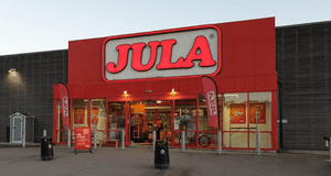 Entrén till Jula-butik med stort rött skyltparti och logotyp ovanför ingången. Glasdörrar leder in till butiken och öppettider visas till vänster på fasaden.