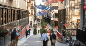 Fotgängare på en sluttande gågata i Stockholm med "MOOD Stockholm"-skylten och färgglada segel uppspända mellan fasaderna.