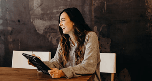 En medelhög inomhusbild visar en leende kvinna med långt, mörkt hår som sitter vid ett robust träbord. Hon är klädd i en mjuk, ljusgrå stickad tröja och tittar bort åt höger med ett brett, genuint leende. Kvinnan håller en svart surfplatta eller en bok me