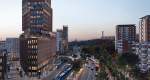 En arkitektonisk visualisering av en modern skyskrapa i tegel som reser sig över en livlig stadsmiljö i skymning. Vid byggnadens fot syns spårvagnar, bussar och biltrafik, omgivna av lummiga trädalléer och intilliggande bostadshus.