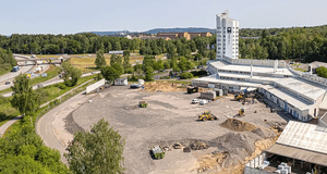 En översiktsbild av en vit låg byggnad, med ett tillhörande högre torn. I mitten av bilden syns en grusplan och en mindre gul traktor som schaktar grus.