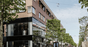 En solig gågata i en stadsmiljö med flervåningshus i tegel, stora butiksfönster i glas och en rad av gröna träd längs trottoaren.