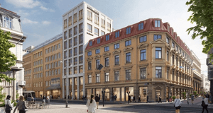 En stadsgata med flera byggnader i klassisk och modern stil. I förgrunden syns människor som promenerar och några sitter vid uteservering. Till höger står en större hörnbyggnad i ljus tegel med rödmålat tak och många fönster i rad. Bredvid finns en högre,