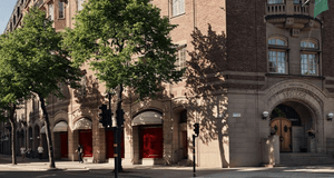 En exteriörbild tagen på gatunivå visar en del av en ståtlig, äldre hörnbyggnad i rött tegel med en bas i ljusare natursten. Byggnaden har arkitektoniska detaljer som rundbågade fönster och en markerad entré i hörnet. Entrén pryds av en inskription ovanfö