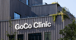 Exteriör av GoCo Clinic. Den moderna byggnaden har en fasad av lodräta träribbor och stora vita bokstäver som bildar namnet. På takterrasserna syns gröna växter och hängande klätterväxter mot en klarblå himmel.