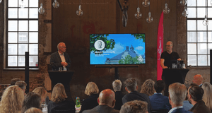 En presentation pågår i en industrilokal med stora fönster och många hängande glödlampor i taket. Två män står vid varsin talarpod och talar inför en sittande publik. Mellan dem syns en stor skärm med en presentation som visar ett porträttfoto, namnet And