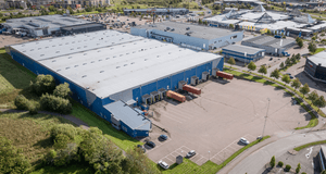 Flygfoto över ett stort industri- eller logistikområde dominerat av en blå och grå lagerbyggnad. Byggnaden har en stor, ljusgrå takyta och en lång blå fasad med en rad lastportar. Framför lastportarna finns en asfalterad lastgård där flera röda och vita t
