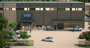 En modern brun byggnad med skyltar för Lunds universitet och Wihlborgs på fasaden. Utanför entrén syns människor som går eller cyklar, samt flera parkerade bilar och en liten lastbil. Byggnaden har stora fönster på andra våningen och omges av ett staket o