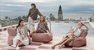 Fyra kvinnor poserar i stilrena kläder i jordnära toner på en takterrass med pool. De sitter och står vid rosa sittpuffar på ett schackrutigt golv, med havet och en palm i bakgrunden under en molnig himmel.