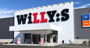 Exteriör av en Willys-butik med den klassiska logotypen på en vit fasad ovanför entrén. Till höger syns skyltar för Svenska Spel, DHL och uttagsautomat, och framför ingången finns parkerade cyklar under en klarblå himmel.