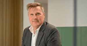Porträtt av en leende man i grå kavaj och vit skjorta, fotograferad i en ljus kontorsmiljö.