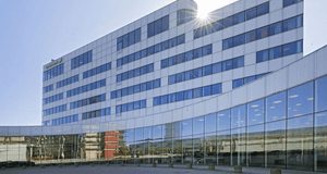 Modern, böjd kontorsbyggnad i Stockholm klädd med vita paneler och horisontella rader av blåfärgade fönster. Entrén är en stor, rundad glasfasad som reflekterar omgivningen. Logotypen "Ericsson" är synlig på den övre delen av fasaden, och starkt solsken s