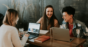 Tre glada personer sitter runt ett bord och samarbetar med bärbara datorer i en modern cafémiljö
