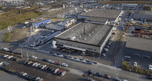 Flygfoto över ett modernt industri- eller handelsområde. I mitten syns en stor grå byggnad med platt tak, och texten "STRIDSYXAN 7" över huvudingången. Byggnaden är omgiven av stora parkeringsplatser fyllda med bilar, samt andra kommersiella fastigheter o