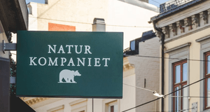 Grön butiksskylt med texten NATUR KOMPANIET och en björnlogotyp som hänger ut från en fasad