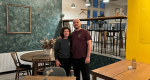 En kvinna med mörkt lockigt hår och en man i en vinröd t-shirt står tillsammans i en restaurang- eller caféinteriör. De står framför en mörkgrön, mönstrad fondvägg med en tom tavelram och ett runt träbord med stolar. Till höger bryter en starkt gul vägg a