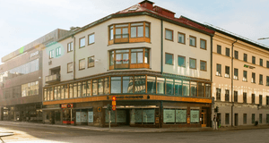 Hörnfastighet i tre våningar med ljus puts, trädetaljer och skyltfönster vid en korsning under en solig dag.