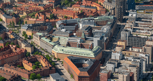 En flygbild över en tätbebyggd stadsmiljö med en unik triangelformad tegelbyggnad i centrum, omgiven av klassiska husrader och moderna höghus under en klarblå himmel.