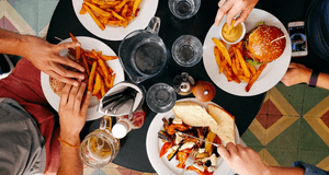 Vy uppifrån på ett runt matbord där fyra personer äter. På tallrikarna finns hamburgare, pommes frites och en grillad grönsakssmörgås. En person doppar pommes i sås, en annan håller i en ölsejdel, och en har en smartphone bredvid sig. Bordet står på ett f