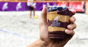 En hand håller upp en genomskinlig bägare med en açaí-bowl från Oakberry. Innehållet är varvat med lila açaí och jordnötssmör, toppat med blåbär. I bakgrunden syns en sandplan där personer spelar sport.