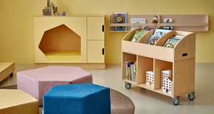 Modern förskolemiljö med trämöbler, en rullbar bokhylla fylld med barnböcker, sittpuffar och lekhörna.