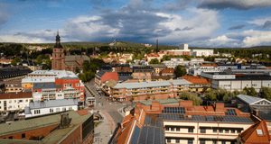 Flygfoto i fågelperspektiv över ett stadslandskap med kyrka, tegelbyggnader med solceller och skogsklädda höjder i bakgrunden under en dramatiskt molnig sky.