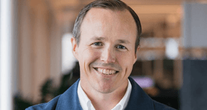 Ett närporträtt av en leende man med kort ljusbrunt hår. Han bär en vit skjorta under en blå, texturerad kavaj eller kofta. Bakgrunden är ljus och mjukt suddig, vilket ger bilden en professionell känsla.