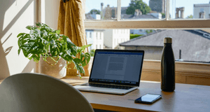 Inomhusbild som visar ett brunt träbord framför ett fönster. En silverfärgad bärbar dator med ett öppet dokument och en svart termosflaska står på bordet. Till vänster står en grön krukväxt. Utanför fönstret syns hustak och ett gammalt kyrktorn i bakgrund