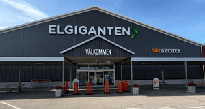 Exteriörbild i ögonhöjd på entrén till ett Elgiganten-varuhus med skyltarna ELGIGANTEN, VÄLKOMMEN och Kronans Apotek under en klarblå sky.