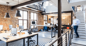 Interiörbild av ett modernt kontorslandskap i industriell stil. Rummet har tegelväggar, stora fönster och synliga stålbalkar. Flera personer arbetar vid ljusa skrivbord, och en man i rörelse går nedför en betongtrappa till höger. Golvet är täckt med en sl