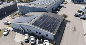 Flygfoto över en industrifastighet med stora solcellspaneler installerade på taken. Byggnaden omges av en parkeringsplats med bilar och ligger i anslutning till ett grönområde.